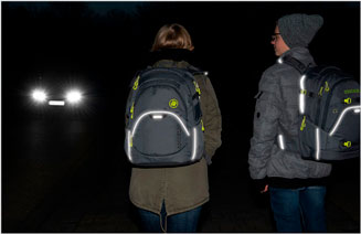 Zwei Kinder in der Nacht mit Schulrucksäcken. Reflektierende Bereiche sichtbar. Auto kommt entgegen.